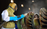 man in protective gear sandblasting a giant metal drill