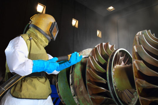 man in protective gear sandblasting a giant metal drill