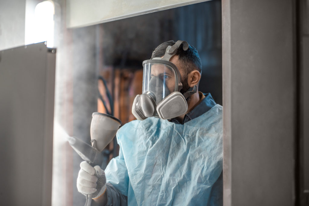 a man in a ventilated face mask and blue protective clothing spraying powder coating onto a surface